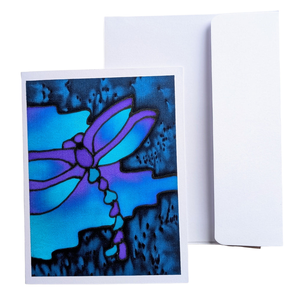 A hand painted pure silk original art  greeting card with an abstract stained-glass style dragonfly design in blue, purple, and black, shown in front of a plain white envelope on a white background.