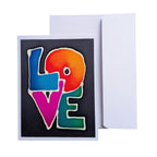 A hand painted pure silk original art  greeting card with the word LOVE spelled out in large, colorful letters over a dark background, shown with a plain white envelope behind it on a white studio background.