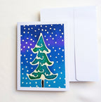 hand painted silk original art greeting card with a hand-painted green Christmas tree on a blue and purple gradient snowy background, shown next to a plain white envelope 